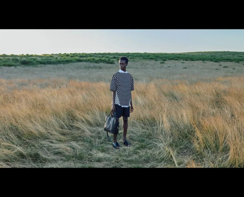 Young Black adult stands in golden field wearing striped shirt and shorts holding dark bag. Grassy meadow stretches to the horizon under clear sky.