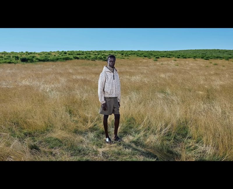 Model standing in a vast dry meadow wearing a light patterned hooded jacket and dark shorts under a clear blue sky.