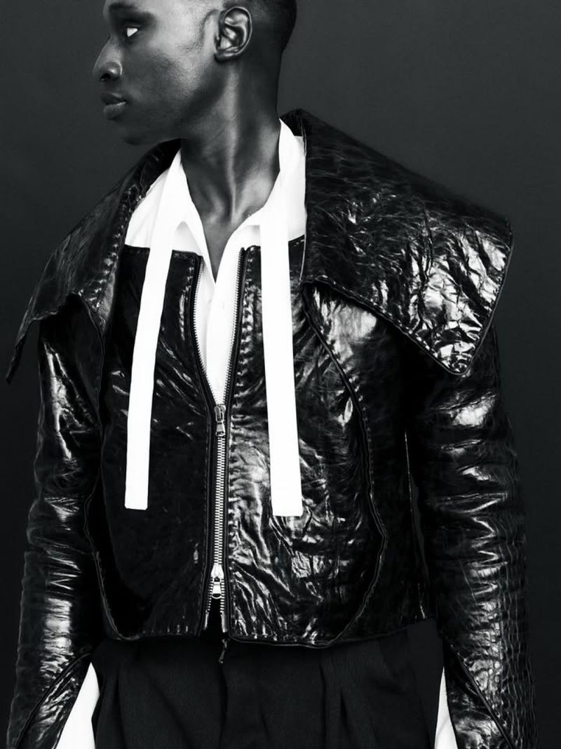 Black and white portrait of a person in a shiny black jacket with a zipper and white straps, and a white shirt against a dark background.