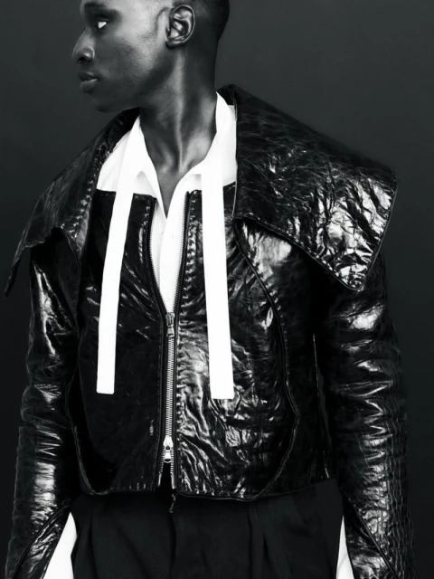 Black and white portrait of a person in a shiny black jacket with a zipper and white straps, and a white shirt against a dark background.