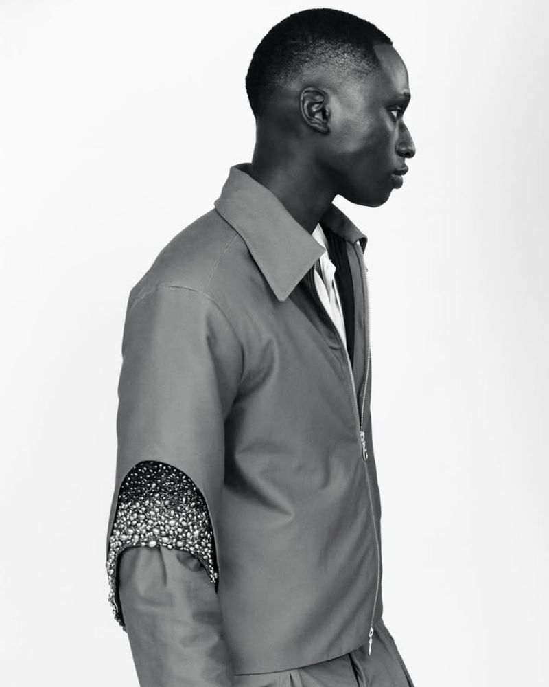 Profile of an adult male in a grey zip-up jacket with arm cut-out and bead detail standing in a photo studio with a white backdrop.
