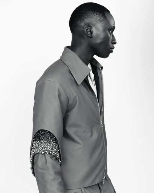 Profile of an adult male in a grey zip-up jacket with arm cut-out and bead detail standing in a photo studio with a white backdrop.