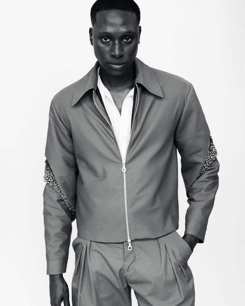 Black male model poses in gray zip-up jacket with embellished sleeves and matching pants in a neutral studio setting.