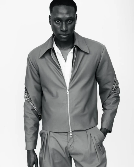 Black male model poses in gray zip-up jacket with embellished sleeves and matching pants in a neutral studio setting.