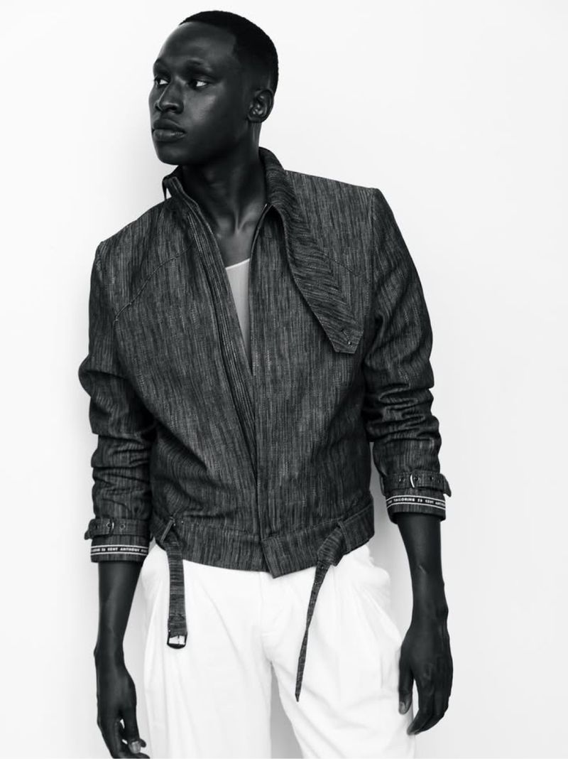 Monochrome fashion shot of an African-American male model wearing a textured jacket and white pants against a bright, neutral background.