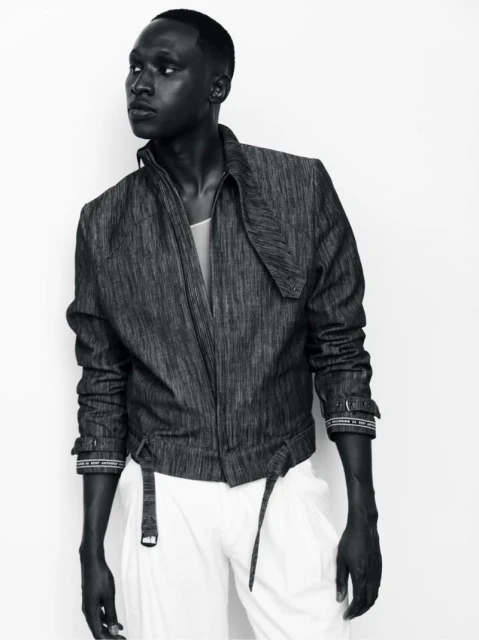 Monochrome fashion shot of an African-American male model wearing a textured jacket and white pants against a bright, neutral background.