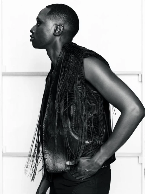 Portrait of a Black male model with a shaved head, wearing a sleeveless fringed leather vest, hand in pocket, in a photo studio.