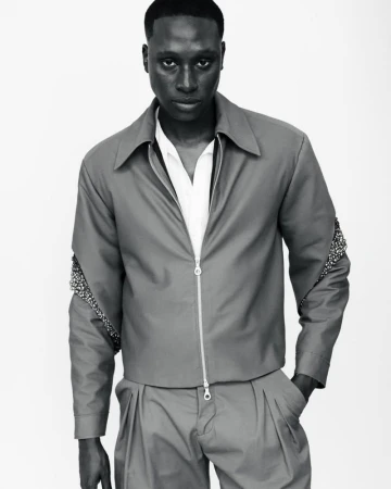 Black and white studio portrait of a male model wearing a zipped jacket and pleated trousers against a seamless white background.