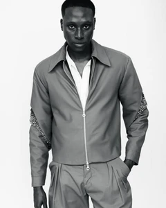 Black and white studio portrait of a male model wearing a zipped jacket and pleated trousers against a seamless white background.