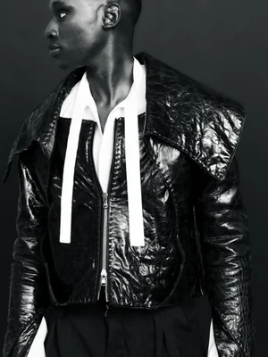 Close-up portrait of a Black male model wearing a glossy black textured jacket over a white collared shirt against a black background.