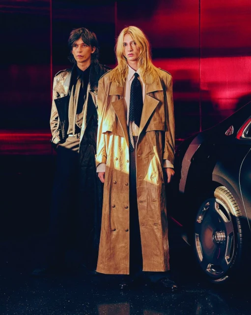 Two people standing next to a car, one wearing a beige trench coat and the other a silver jacket, set against a red background.