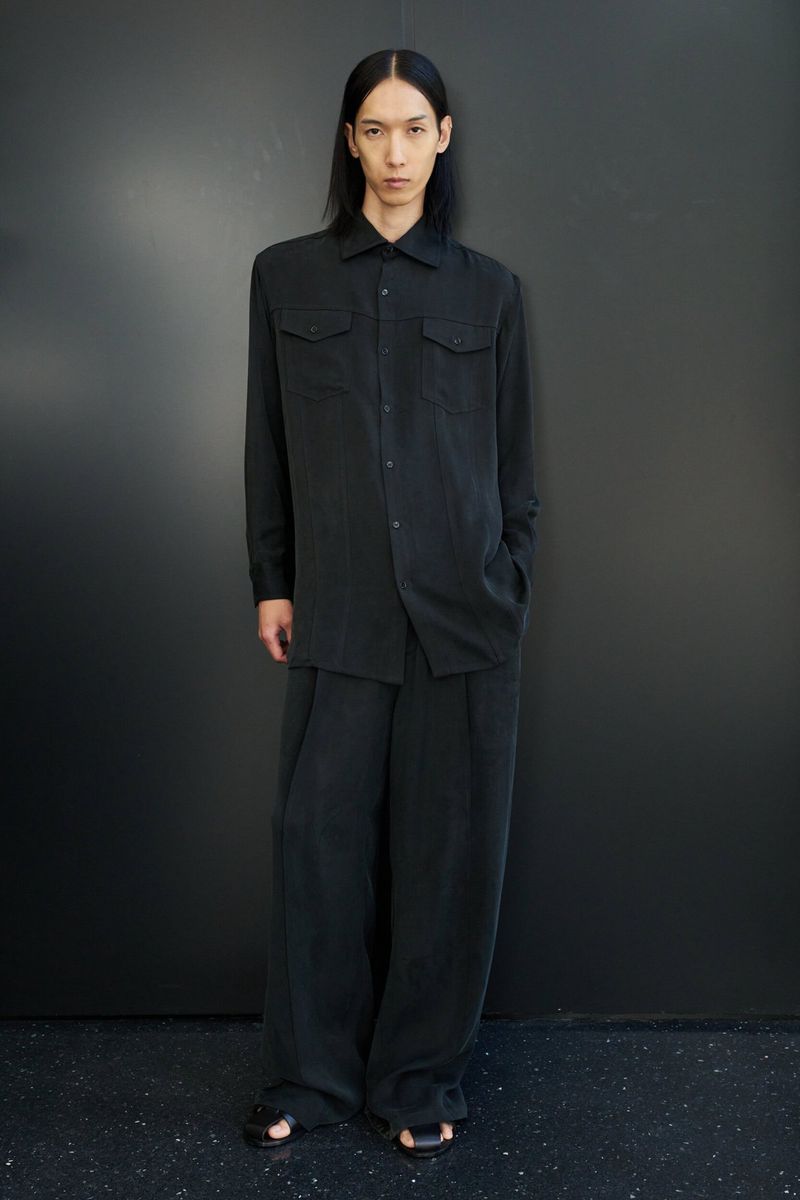 A single adult male poses in black shirt and pants against a dark gray wall, looking at the camera, in a full-length studio shot.