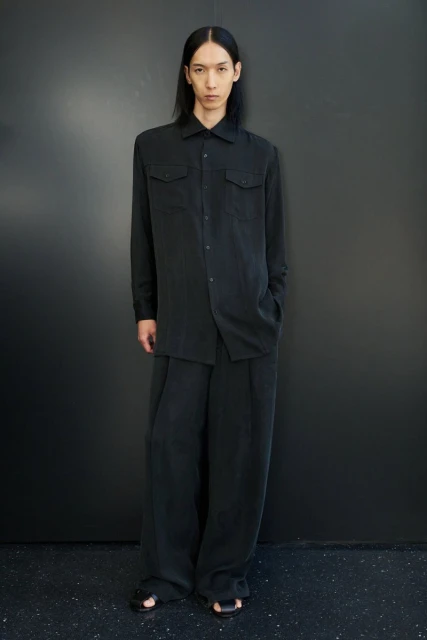 A single adult male poses in black shirt and pants against a dark gray wall, looking at the camera, in a full-length studio shot.