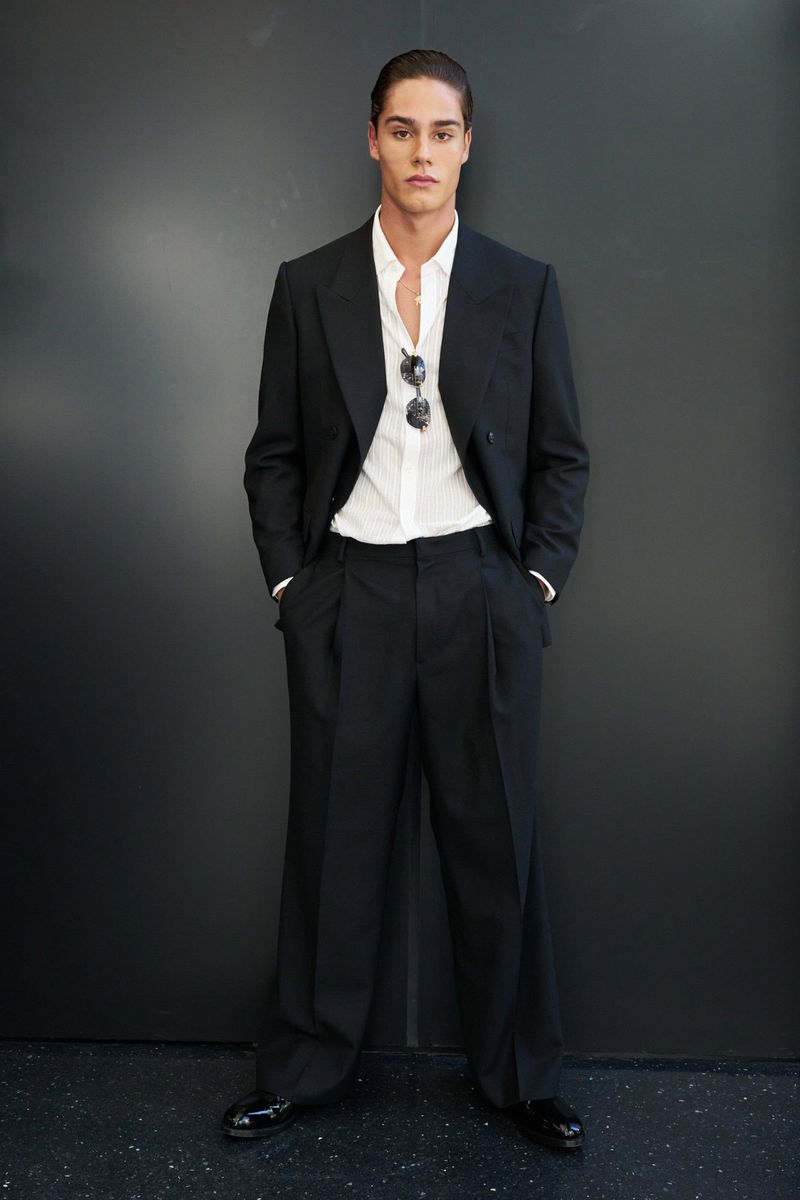 Full-length shot of a young adult man in a black suit and white shirt, standing indoors against a dark gray wall with sunglasses.