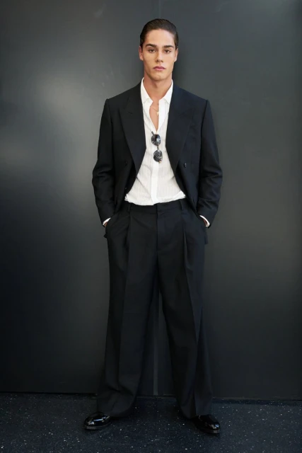 Full-length shot of a young adult man in a black suit and white shirt, standing indoors against a dark gray wall with sunglasses.