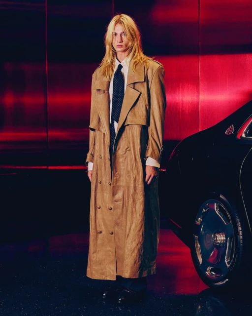 Fashion shot of a blonde adult wearing a trench coat standing next to a car against a dark red background in an indoor setting.