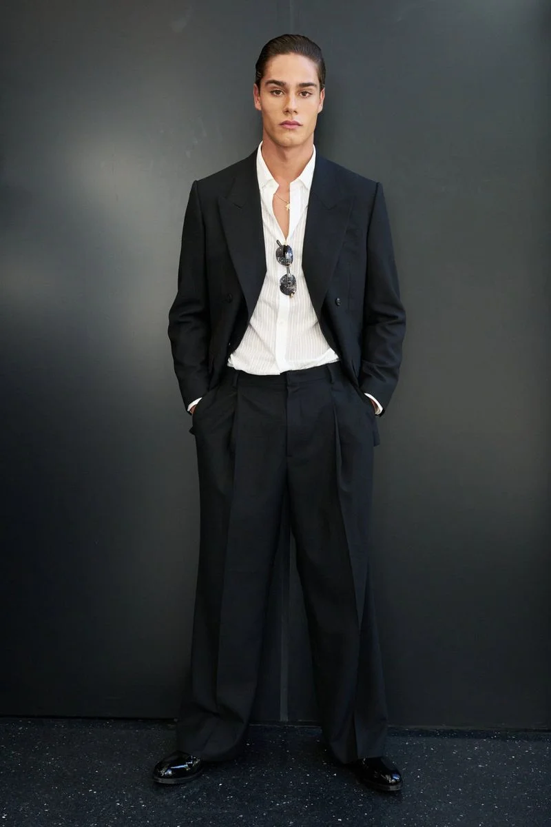 Model standing against a dark wall wearing a black suit and white shirt with sunglasses tucked into the collar