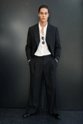 Model standing against a dark wall wearing a black suit and white shirt with sunglasses tucked into the collar