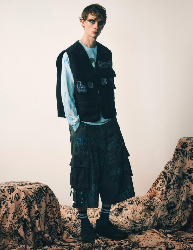 Male model wearing a black vest, patterned t-shirt, and cropped pants, posing on a patterned fabric in a studio setting. Fashion editorial.