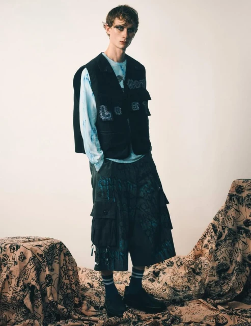 Male model wearing a black vest, patterned t-shirt, and cropped pants, posing on a patterned fabric in a studio setting. Fashion editorial.