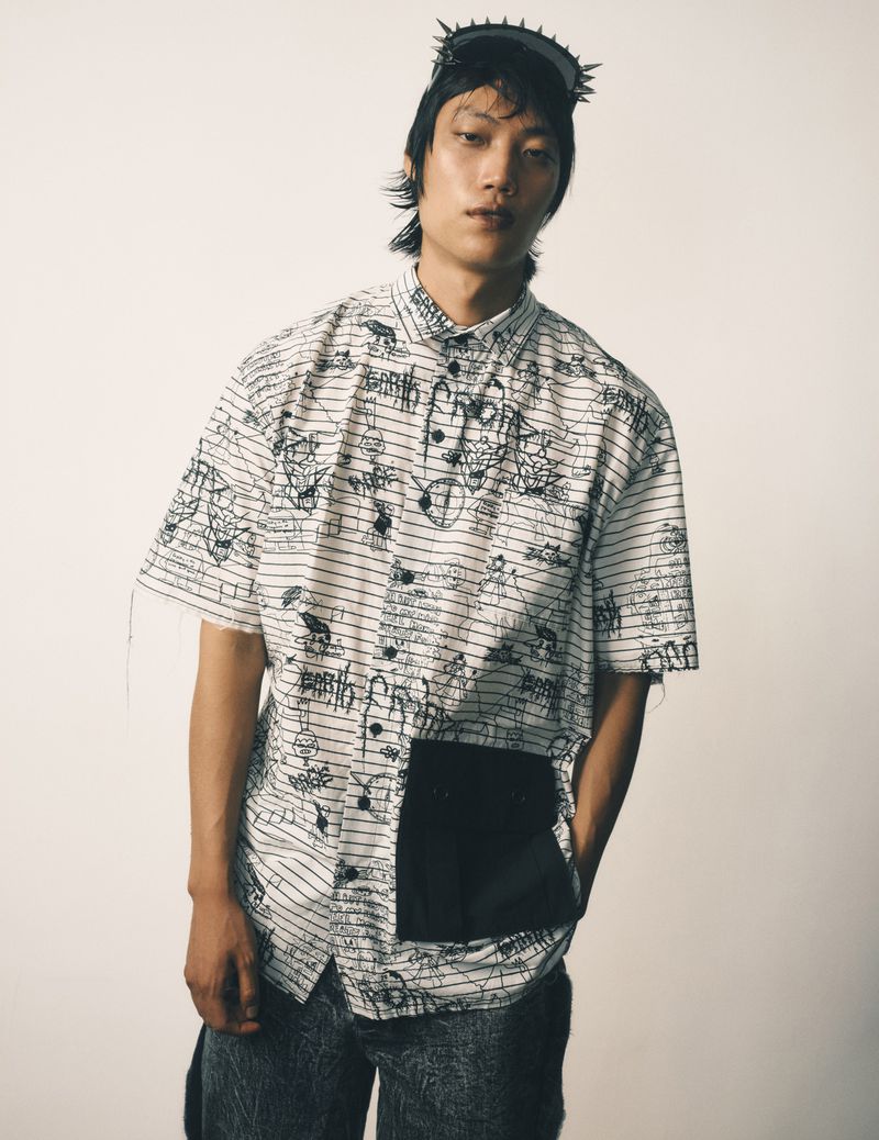 Man wearing graphic shirt, black headdress, and marble-wash pants poses in a studio against a cream-colored background. Fashion shot.