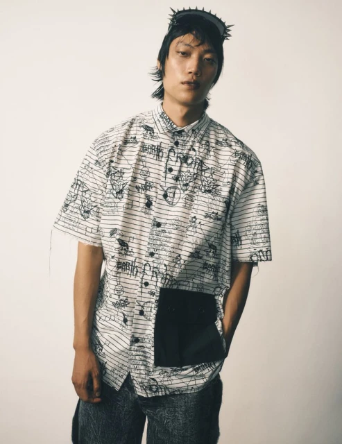 Man wearing graphic shirt, black headdress, and marble-wash pants poses in a studio against a cream-colored background. Fashion shot.