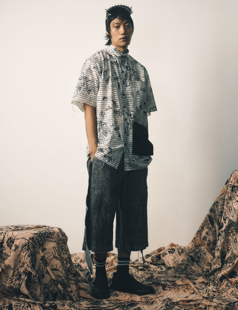 A model poses in an open button-down shirt with dark shorts against a light background with a floral-patterned cloth in the foreground.