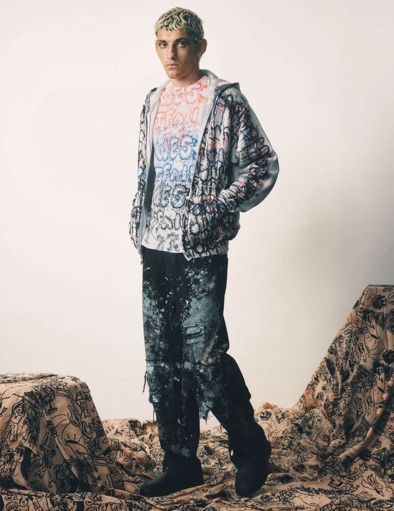 A young adult male poses in a full-length shot, wearing a graffiti-style outfit, against a neutral backdrop with patterned fabric below.