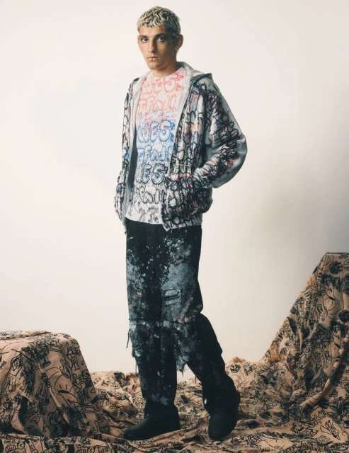 A young adult male poses in a full-length shot, wearing a graffiti-style outfit, against a neutral backdrop with patterned fabric below.