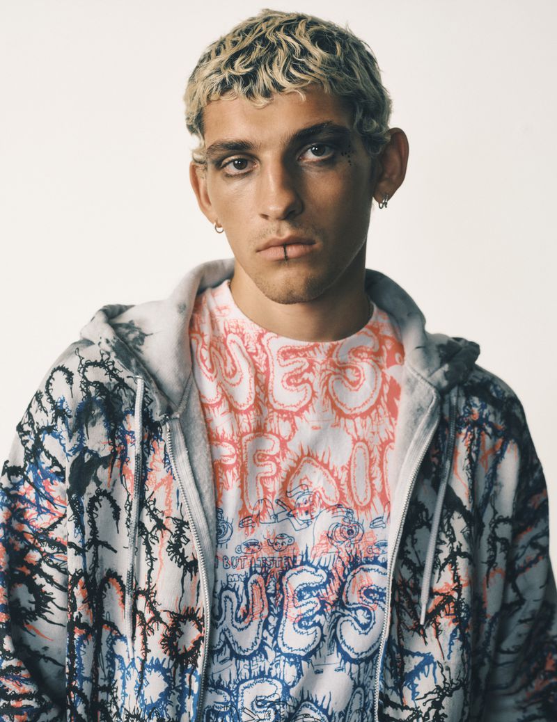 Close-up of a young adult male with blonde hair and black eyeliner, wearing a graphic t-shirt and zip-up hoodie against a plain background.