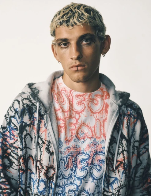 Close-up of a young adult male with blonde hair and black eyeliner, wearing a graphic t-shirt and zip-up hoodie against a plain background.