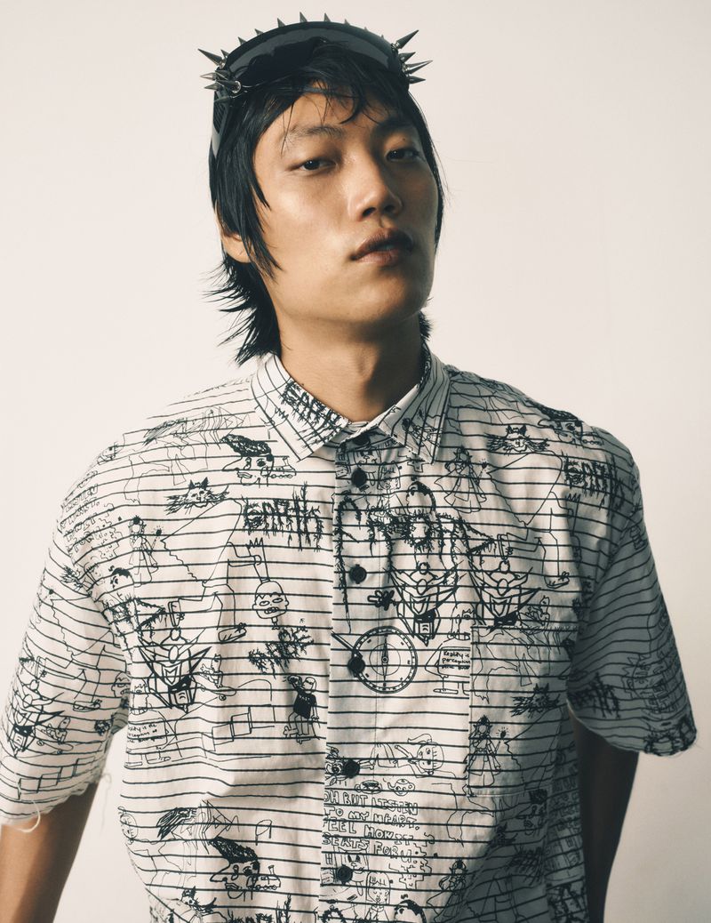 Asian male model poses indoors wearing sunglasses with spikes and a white button-down shirt with black line art and doodles.