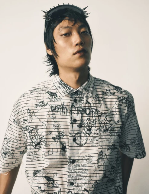 Asian male model poses indoors wearing sunglasses with spikes and a white button-down shirt with black line art and doodles.