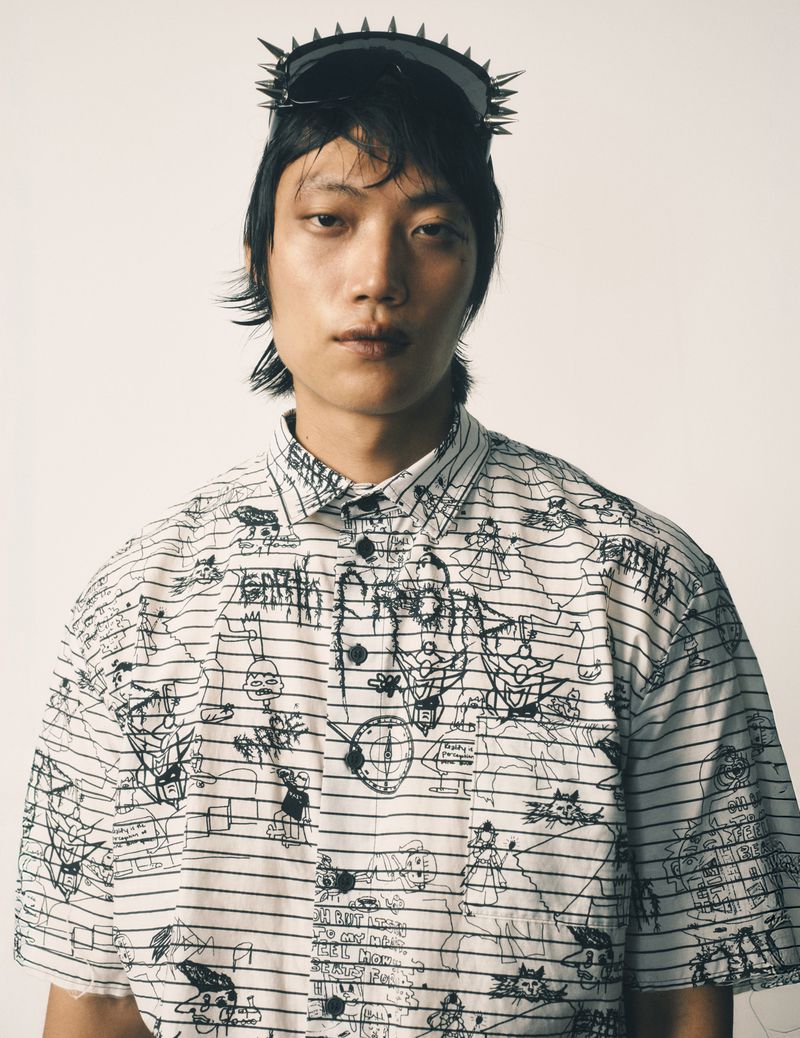 Portrait of a young adult with spiky sunglasses and a patterned button-down shirt, posing against a stark white backdrop.