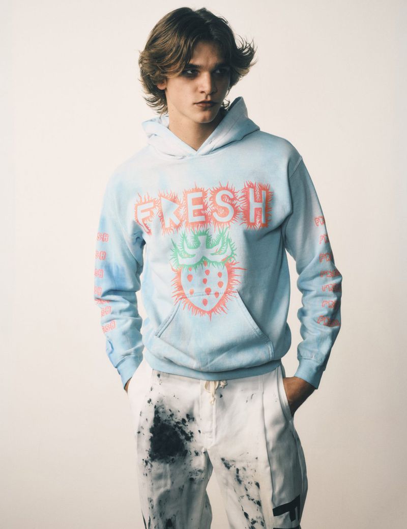Young adult poses indoors wearing a light blue hoodie with a strawberry graphic and paint-splattered white trousers in a studio shot.