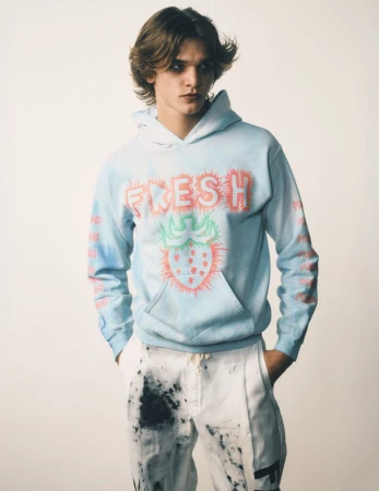 Young adult poses indoors wearing a light blue hoodie with a strawberry graphic and paint-splattered white trousers in a studio shot.
