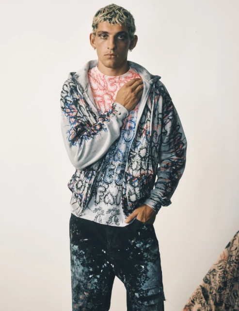 Adult male with bleached hair wearing a graphic hoodie and paint-splattered pants poses in front of a light background in a studio shot.