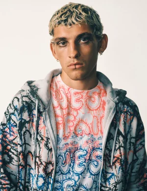 Portrait of a young male model with bleached curly hair wearing a patterned graphic hoodie against a plain white background