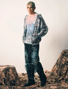 Model standing on patterned fabric in a printed hoodie and dark cargo pants against a plain background