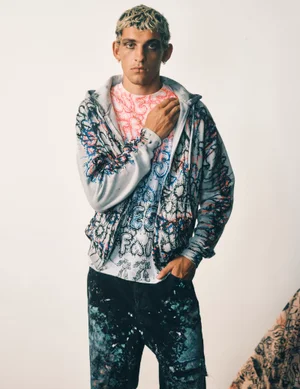 Young man with short curly hair wearing a patterned hoodie and dark trousers against a plain white background.