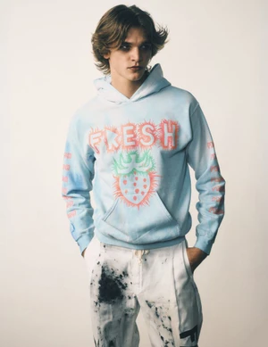 Young man wearing a light blue graphic hoodie and white patterned trousers against a seamless white background.