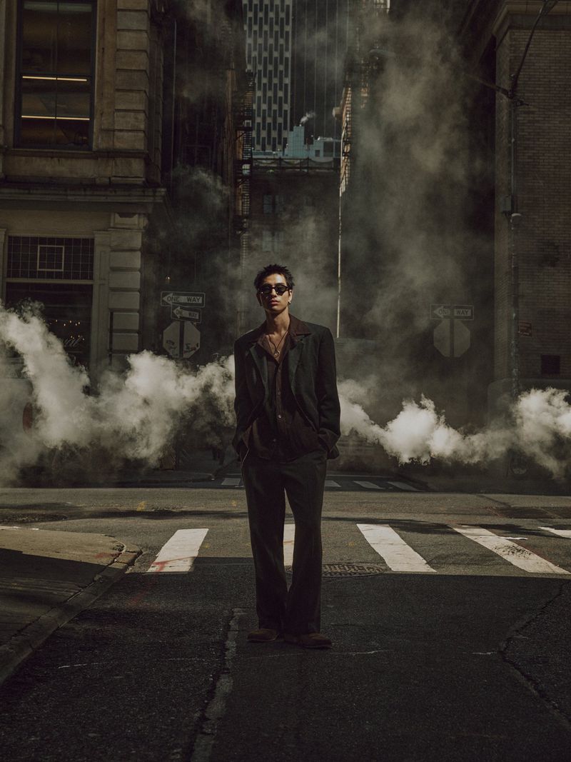 Man standing on a city street amid steam rising from a manhole cover wearing a dark suit