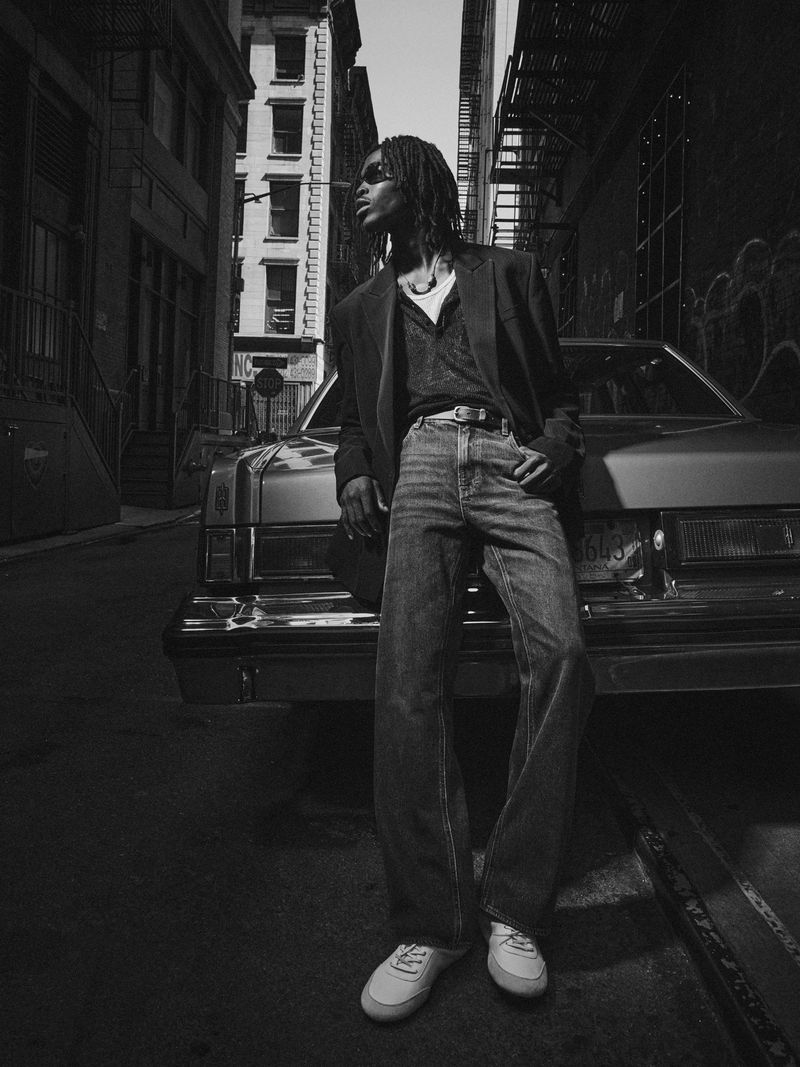 A black and white shot of a Black male model in a suit jacket leaning against an old car in an alley with tall buildings.