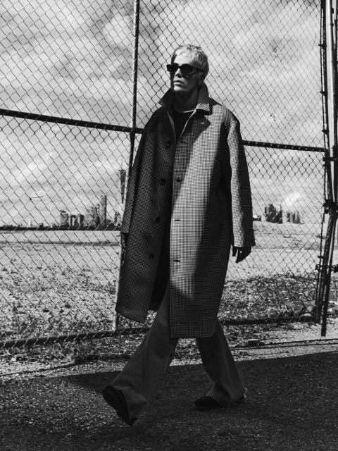 Monochrome portrait of an adult male in sunglasses and an overcoat walking in an outdoor urban setting, against a chain link fence.