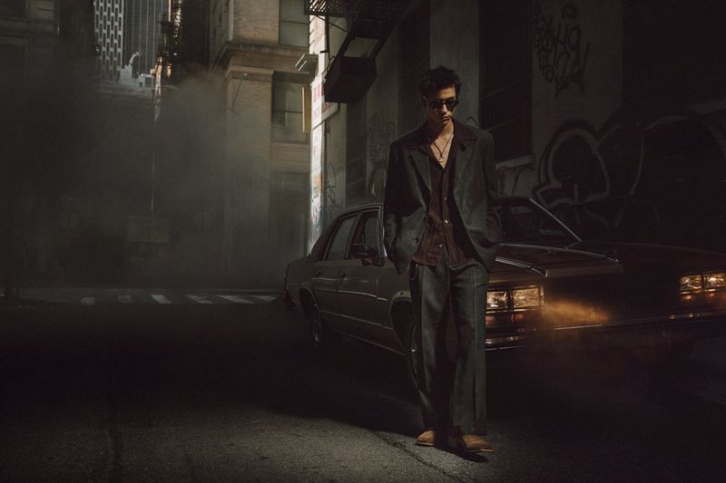 Stylish man in a suit and sunglasses stands near a classic car on a city street with smoke and urban architecture in the background.