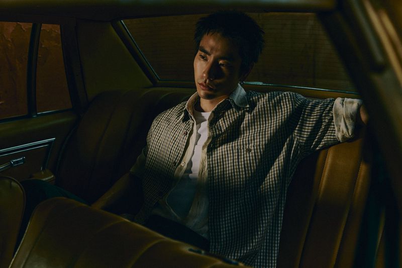 Portrait of a pensive adult man in a plaid shirt sitting in the back seat of a vintage car in a dimly lit, brown interior.