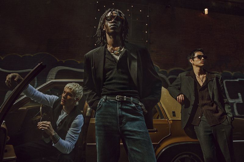 Three stylish adult men pose in front of an old gold car and graffitied wall in an urban setting, wearing fashionable casual-formal clothing.