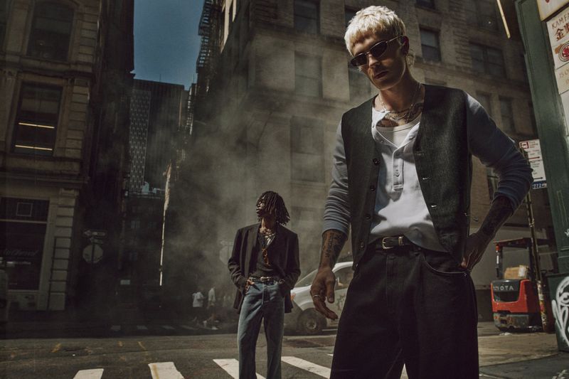 Two men in fashionable attire stand in a city street with tall buildings and steam, bathed in warm sunlight creating shadows and highlights.