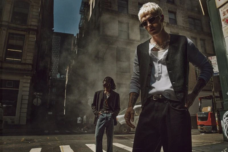 Two men in fashionable attire stand in a city street with tall buildings and steam, bathed in warm sunlight creating shadows and highlights.