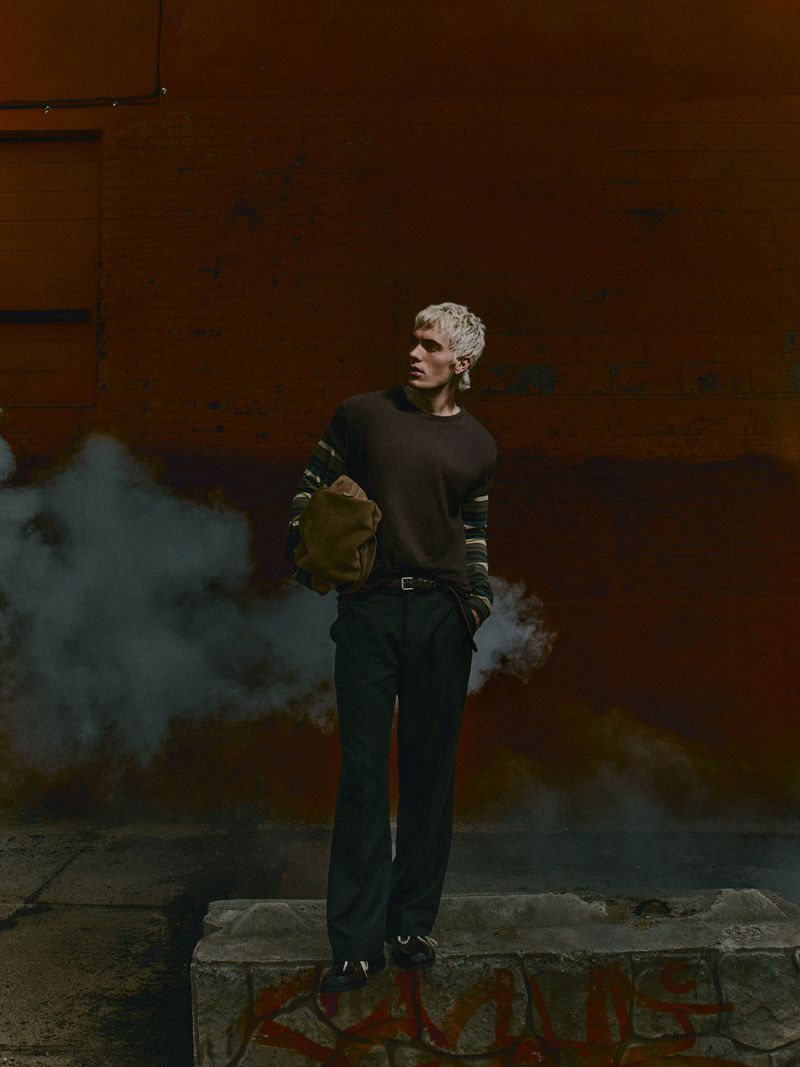 Full-length portrait of an adult male with platinum blonde hair holding a bag, standing in front of a brown concrete wall with smoke.